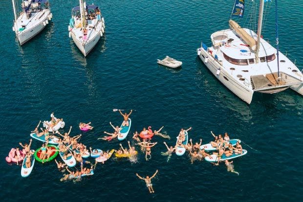 Photo of a large group of people floating on inflatables in turquoise waters of Epidavros Greece with sailing yachts around them.